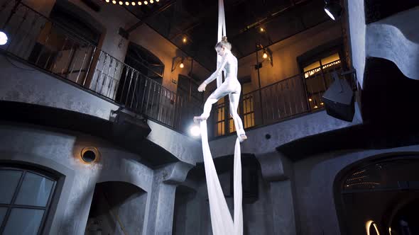 Female aerial acrobat doing a midair split on silks in a club venue.