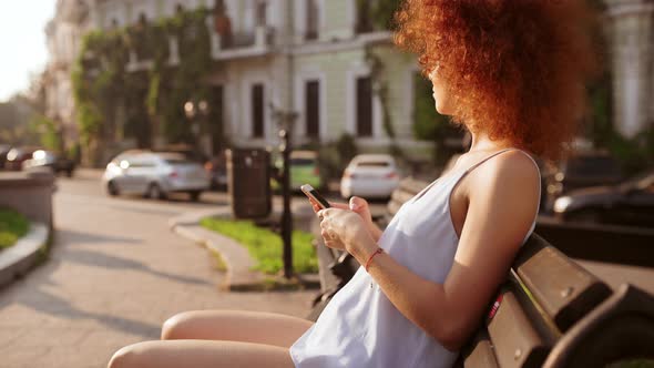 Attractive Readhead Girl Resting Looking at Phone Sitting on Bench Outside