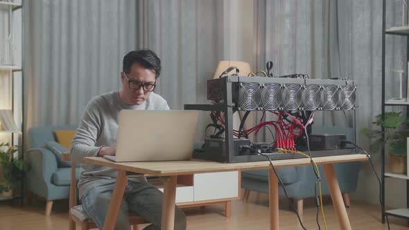 Serious Asian Man Wearing Glasses Using Laptop And Mining Rig For Mining Cryptocurrency