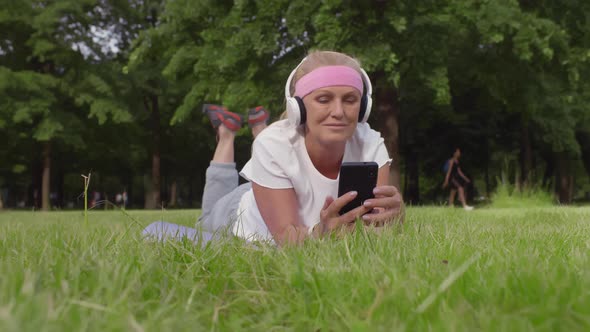 Senior Woman Relaxing on Grass in Headphones Using Smartphone in Park