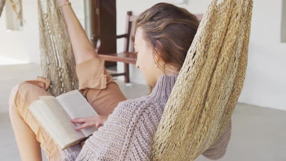 Smiling caucasian woman lying in hammock reading book on terrace