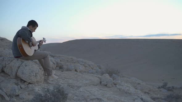 Young adult sits and plays the guitar with desert scenary