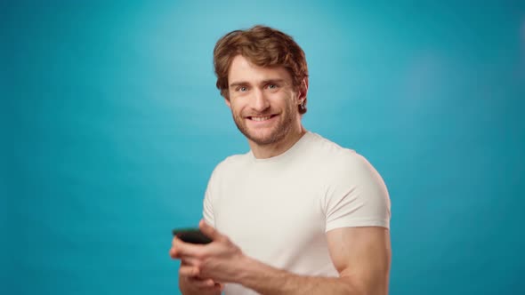 Young Man Looks at Cell Phone Screen with Joy Recieved Good News Against Blue Background