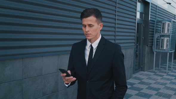 Side View of Confident Businessman in Suit Walking with Phone. Man Looking Into Smartphone Outdoors