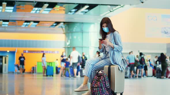 Woman in Mask, Sitting on a Suitcase, Luggage, Against Background of Air Passengers Crowd