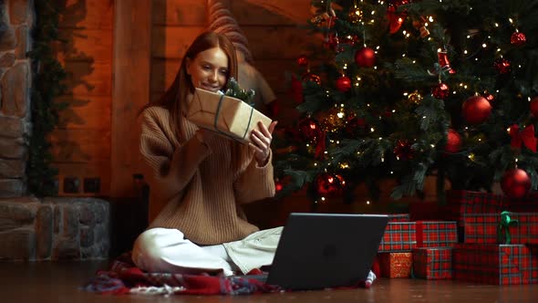 Happy Young Woman Holding Gift Box While Having Video Call on Laptop Sitting Near Christmas Tree.