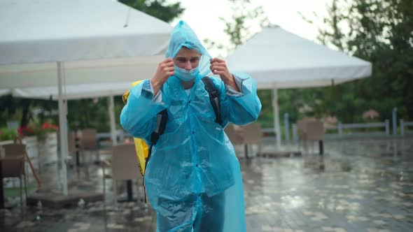 Front View Portrait of Young Delivery Man in Coronavirus Face Mask and Rain Coat Looking Around