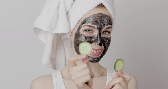 Happy Girl with Towel on Head and Black Facial Mask Posing with Two Cucumber Pieces in Hands