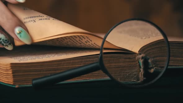 Girl Turns the Page of an Old Book Next to a Magnifying Glass