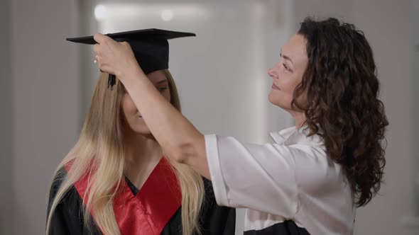 Smiling Mother Putting Graduation Cap Tassel Right to Left on Cap of Happy Daughter Standing Indoors