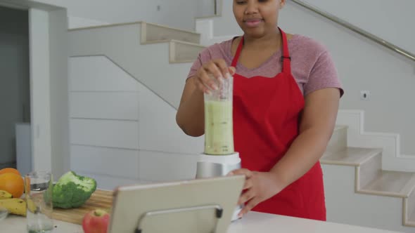 Happy african american plus size woman wearing apron, preparing health drink in kitchen