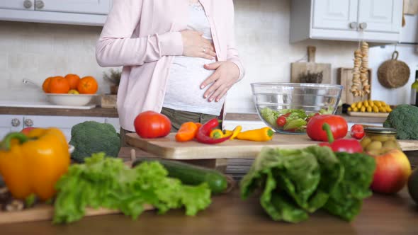 Closeup Of Pregnant Woman Cherishing Her Baby In Belly While Cooking Salad On Kitchen.