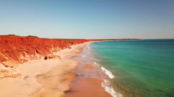 Drone flies beside red cliffs on sandy beach with turquoise blue water, waves break below