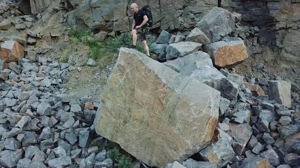 A man in a black t-shirt and a backpack comes to the edge of a large stone and raises his hands up.