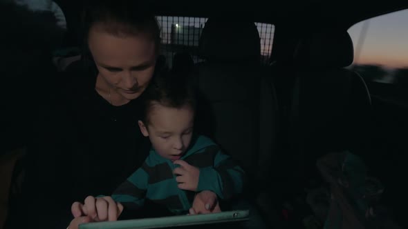 View of Woman with Son in the Dark Riding in Car on Back Seat, Watching Computer Tablet