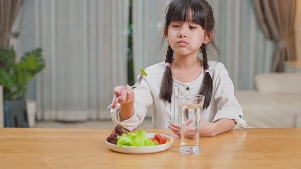 Asian young little girl child eat green vegetable salad in plate on dinner table for health. alt