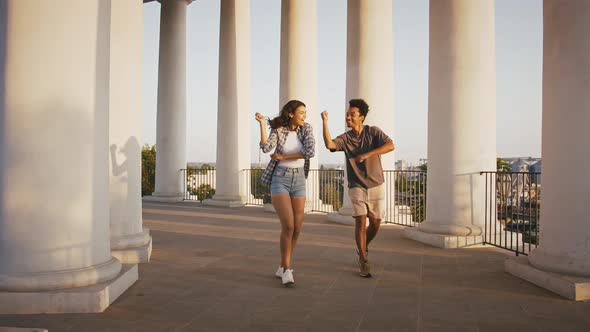 Darkskinned Pair Smiling and Dancing Walking By Pillared Observation Deck in Park