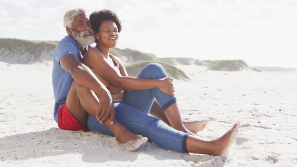 Happy african american couple sitting and embracing on sunny beach