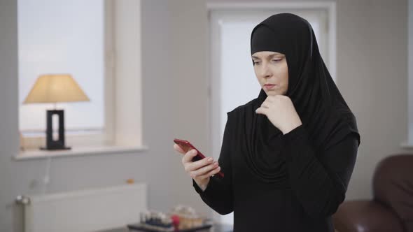 Serious Muslim Woman in Hijab Looking at Smartphone Screen and Scrolling. Young Eastern Lady