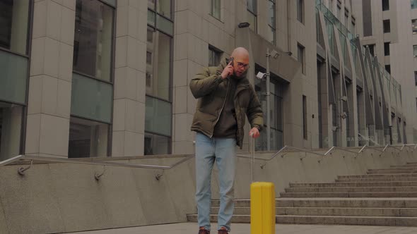 European Man Comes Out with Yellow Suitcase From the Office Center Talking on Phone