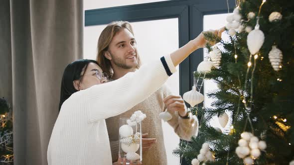 Young Mixed Race Couple are Talking and Smiling Decorating Green Christmas Tree Standing Nearby a