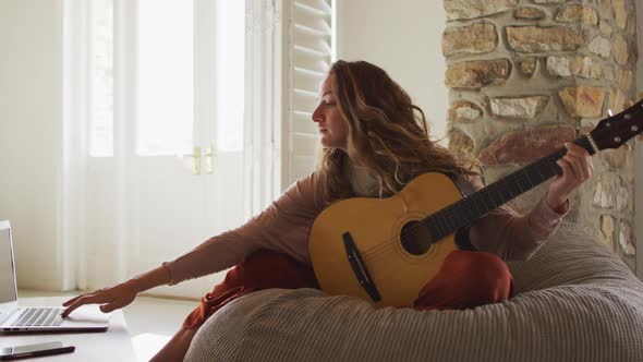 Caucasian woman sitting on beanbag playing acoustic guitar using laptop in sunny cottage living room