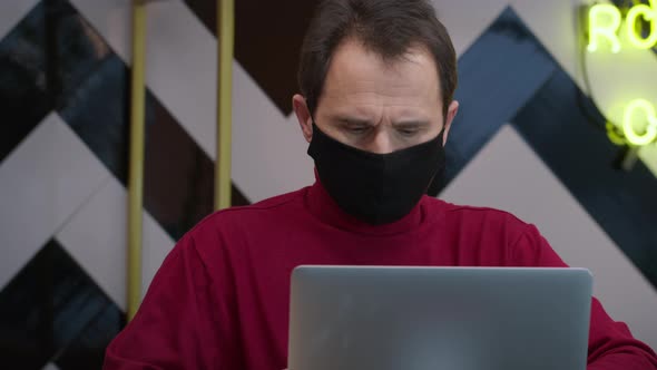 Handsome Middleaged Man in Black Protective Mask Working with Laptop in Cafe