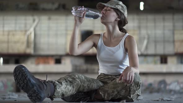 Attractive Young Woman in Military Uniform Drinking Water From the Bottle Sitting on the Floor