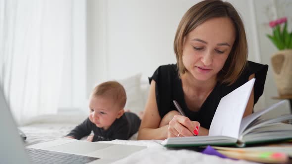 Young Mother Freelancer with Baby Son Sitting on Bed and Trying to Work at Laptop