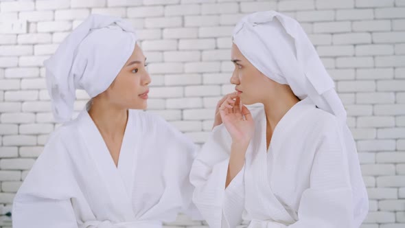 Two Asian girls in white bathrobes with towels on heads talking in living room.