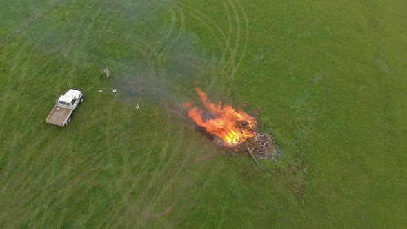 Aerial birds eye view shot flying up above a farmer and his truck by a large bonfire on a farm paddo