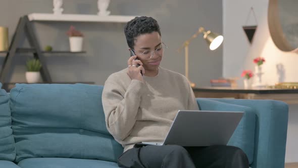 African Woman with Laptop Talking on Smartphone on Sofa