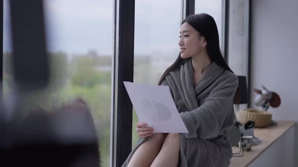 Happy Smiling Asian Female Freelancer in Bathrobe Looking Out the Window Sitting on Windowsill in