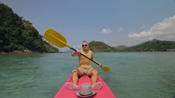 Man with Sunglasses and Hat Rows Pink Plastic Canoe Along Sea Against Green Hilly Islands with Wild