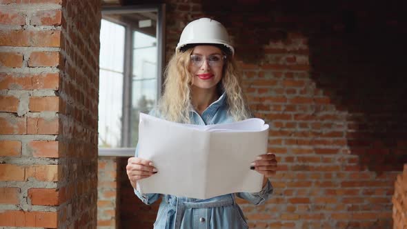 A Young Woman in a White Work Helmet and Denim Clothes and Goggles Stands at the Construction Site