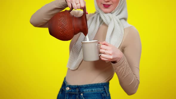 Unrecognizable Young Rural Woman Pouring Milk From Clay Pot in Mug Standing at Yellow Background