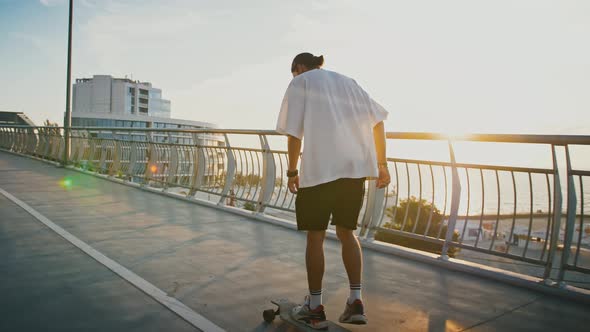 Slow Motion of Millennial Guy Riding Longboard on City Bridge in Evening