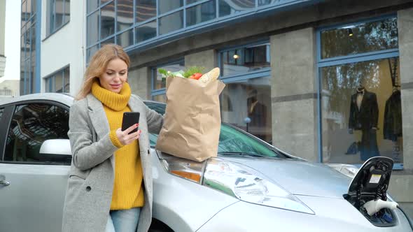 Woman Using Smartphone Near Her Electric Car Groceries Looks Camera