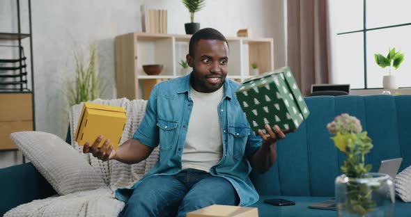 African American Guy Sitting on the Couch at Home and Choosing Between Two Gift Boxes in His Hands