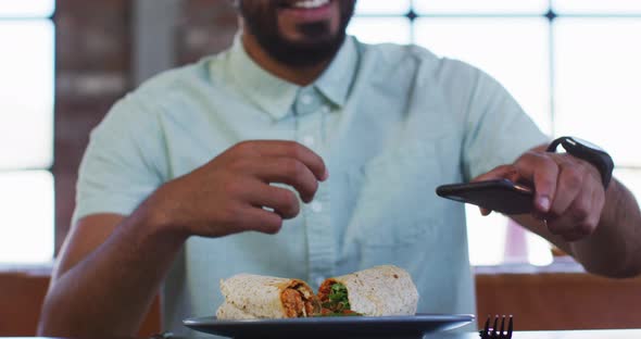 Happy mixed race man sitting in cafe taking picture of his sandwich with smartphone and smiling