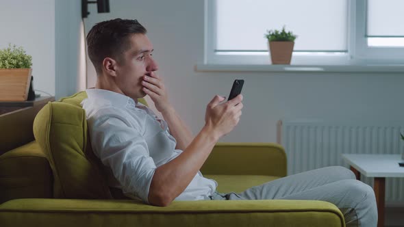 Shocked Man Reacting Message on Smartphone Covers His Mouth in Fright Sitting on Sofa at Home Living