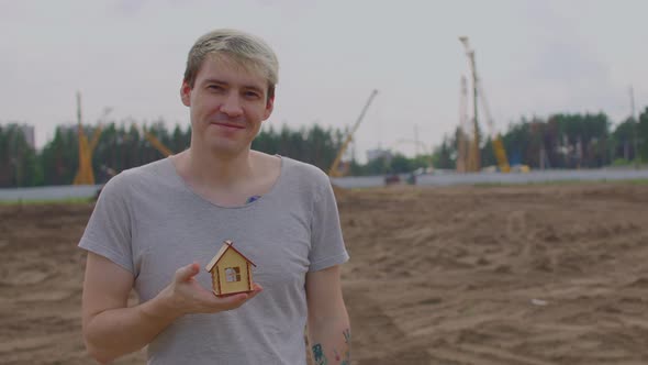 Young Man with Small Wooden House in Hand Stands on Construction Site
