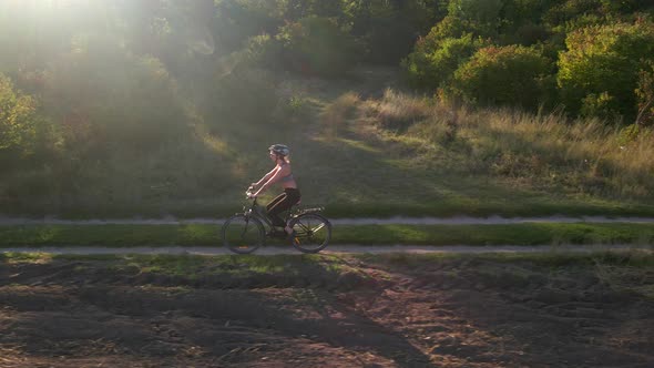 Aerial Slow Motion Shot of Young Sport Woman Rides Bicycle on Countryside Road at Summer Sunset