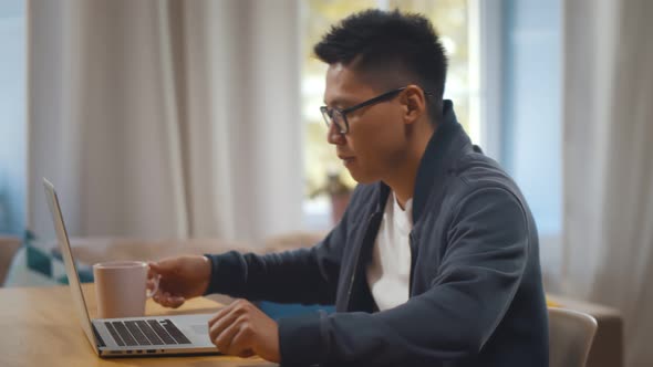 Handsome Young Asian Man Sitting Down at Table with Cup of Coffee Using Laptop Computer at Home