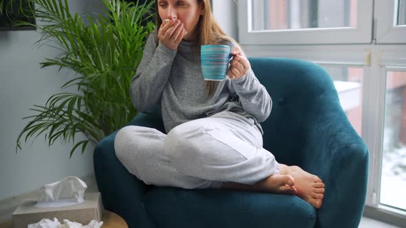 Unhealthy Woman Sits in a Chair Drinks Hot Tea or a Cold Medicine and Sneezes or Blows Her Nose Into