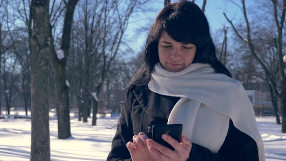 Happy Smiling Beautiful Girl Using Smart Phone. Sunny Day In Snow Covered City Park. Slow Motion