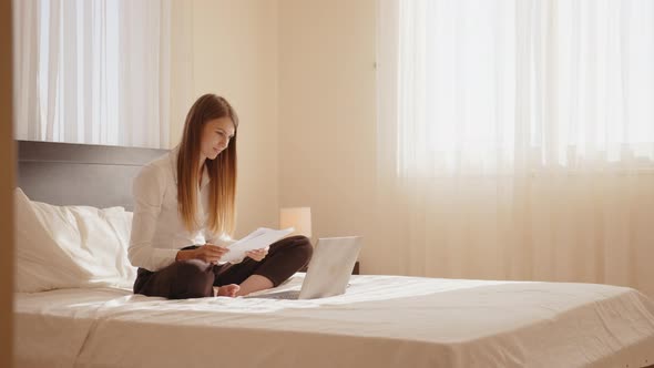 Woman Sitting on Bed with Laptop and Reading Papers at Hotel