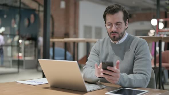 Young Man Using Smartphone While Using Laptop in Office