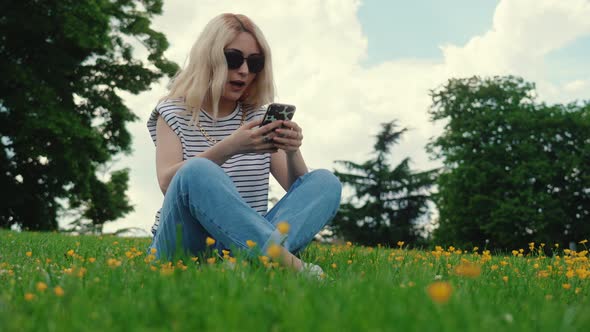 Blond Caucasian Girl Sitting on the Grass and Using Her Mobile Phone Full Shot Outdoors