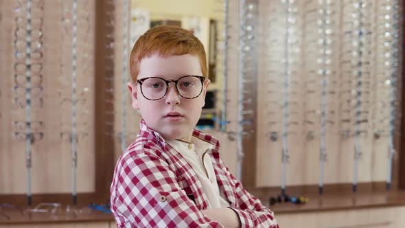 A Redhaired Boy in a Plaid Red and White Shirt Stands with Transparent Glasses for Vision Correction